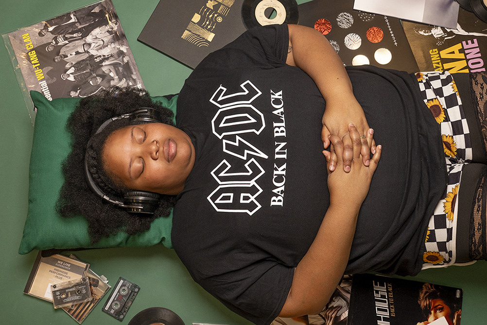Cinnamon laying down with headphones, wearing a black t-shirt that says AC/DC Back in Black and tights with sunflowers and black and white checkers, surrounded by music records and tape cassettes.