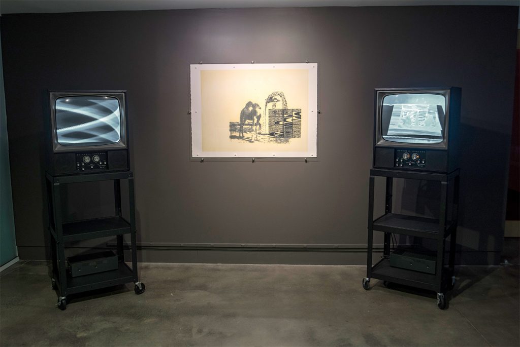 Two CRT monitors displaying black and white abstract imagery with a print hanging between them.