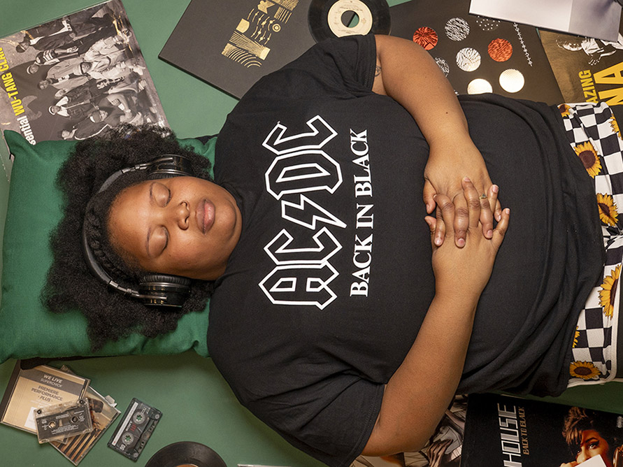Cinnamon laying down with headphones, wearing a black t-shirt that says AC/DC Back in Black and tights with sunflowers and black and white checkers, surrounded by music records and tape cassettes.