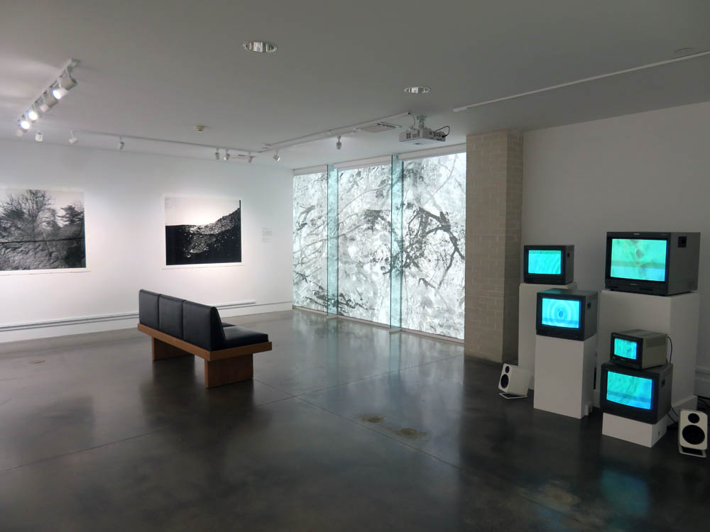 Large gallery space with two photo prints on the far wall; five CRT monitors show a blue video on the right side of the image.