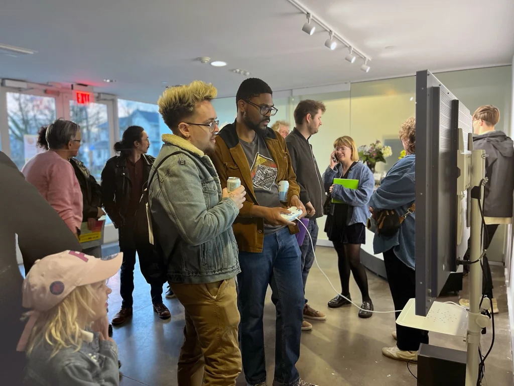 A group of people stand around a TV monitor, one holding a game controller.